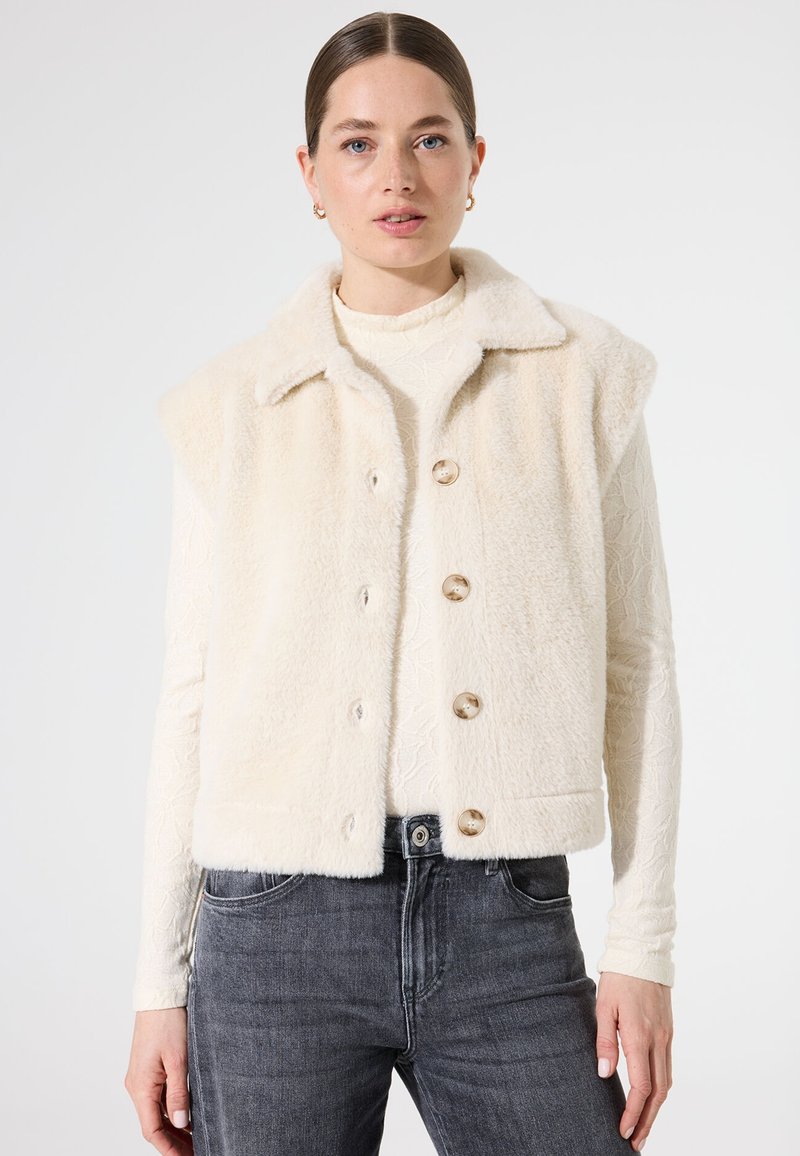 Kunstbont mouwloze vest in crème met een kraaghalslijn, vijf grote ronde knopen en textuurstof. Gecombineerd met grijze jeans.