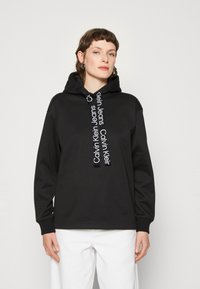 Felpa nera con cappuccio e cordini bianchi con logo. Presenta maniche lunghe, polsini a coste e una vestibilità rilassata. Tessuto liscio, design minimal.