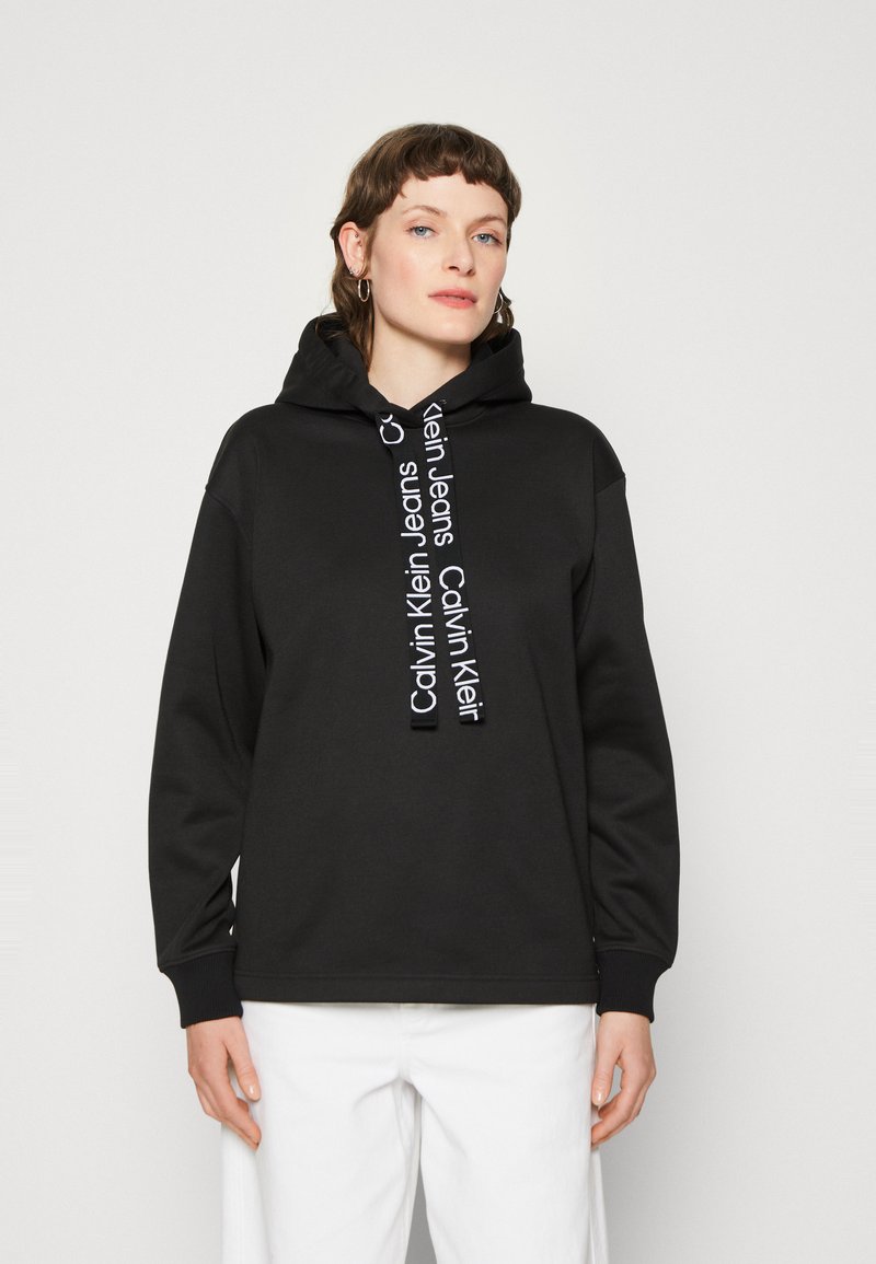 Felpa nera con cappuccio e cordini bianchi con logo. Presenta maniche lunghe, polsini a coste e una vestibilità rilassata. Tessuto liscio, design minimal.