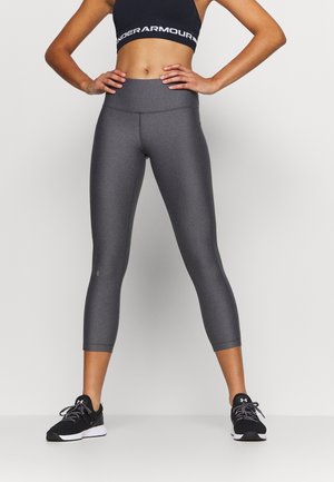 Vrouw draagt een zwarte sportbeha, grijze cropped legging en zwarte sportschoenen, staat met handen in de zij tegen een effen achtergrond.