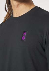 Camiseta de algodón negra con un emblema rectangular que presenta el logo rosa de adidas y un diseño circular en el lado izquierdo cerca del escote.