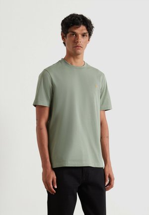 T-shirt in cotone verde menta con scollatura tonda, maniche corte e un piccolo logo dorato sul petto a sinistra. Abbinato a pantaloni neri.