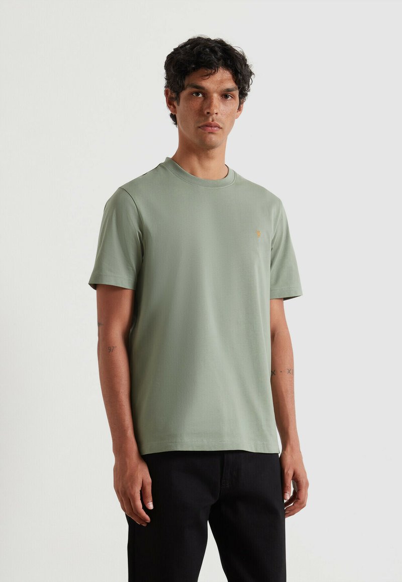 T-shirt in cotone verde menta con scollatura tonda, maniche corte e un piccolo logo dorato sul petto a sinistra. Abbinato a pantaloni neri.