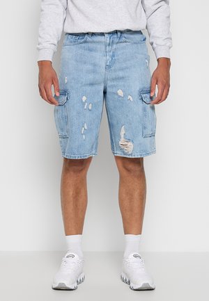 Homme portant un short cargo en denim bleu clair déchiré, des baskets blanches, des chaussettes blanches et une chemise à manches longues gris clair, debout devant un fond uni.