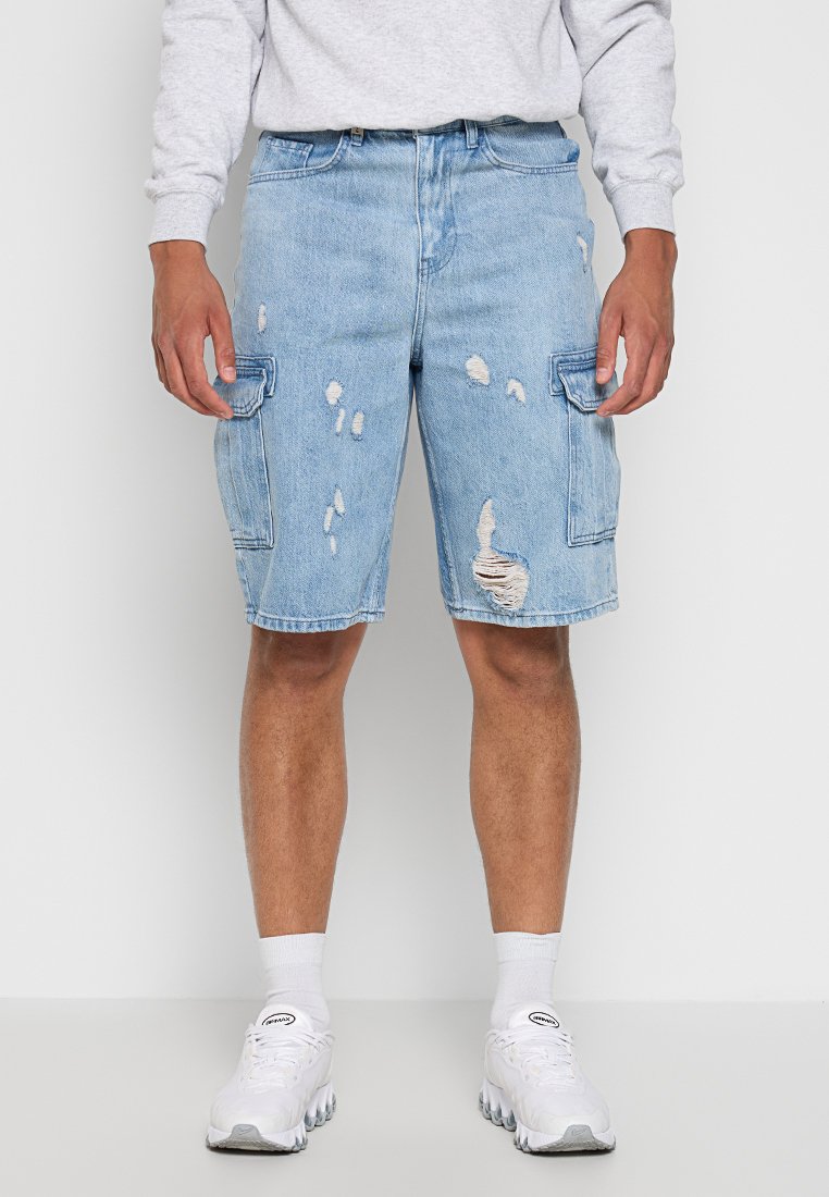 Homme portant un short cargo en denim bleu clair déchiré, des baskets blanches, des chaussettes blanches et une chemise à manches longues gris clair, debout devant un fond uni.