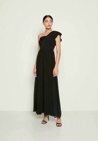 Vestido maxi preto com um ombro de babado, corpete ajustado e saia fluida. O material parece suave e leve. Saltos alto completam o visual.
