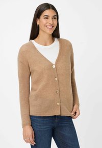 Cardigan beige con scollo a V, cinque bottoni in legno, maniche lunghe e motivo a maglia testurizzato, indossato sopra una maglietta bianca e jeans blu.