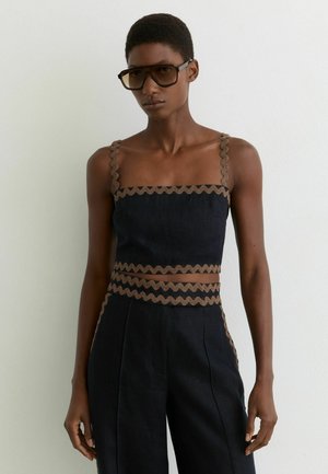 Femeie cu păr scurt purtând ochelari de soare mari și un top negru fără mâneci, cu margini ondulate maro, asortat cu pantaloni negri cu talie înaltă.