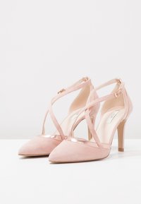 Rosa mockaklackar med spetsiga tår, korsade remmar och metalliska detaljer på ovansidan. Har en stilettklack och en slät insida.