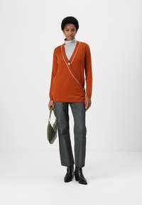 Ein rostfarbener Wickelpullover mit weißem Saum und Knopfdetails, darunter ein grauer Rollkragenpullover, kombiniert mit dunklen, strukturierten Hosen und schwarzen Stiefeletten.