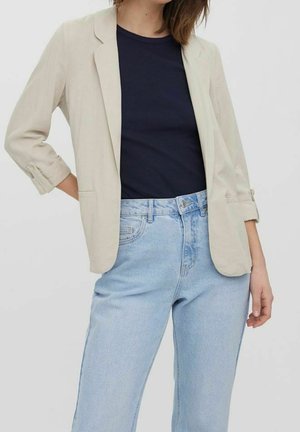 Femme portant un blazer beige aux manches retroussées, un t-shirt bleu marine et un jean taille haute bleu clair, debout avec une main sur la hanche.