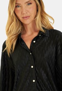 Camisa negra de botones con una tela texturizada y elegante que presenta patrones ondulados, terminada con botones dorados brillantes y un cuello clásico.