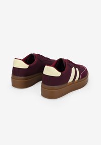 Zapatillas burdeos con material suave, detalles en crema en el talón y dos rayas blancas en el lateral. Suela plana de goma.