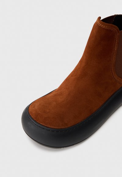 Pruun suede pahkluumoodne saabas, millel on sile must kummitald ja elastsed külgpaneelid, ümarad varbad ja minimaalne õmblusdetail.