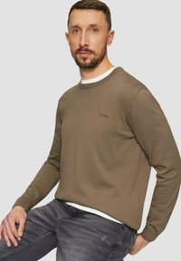 Brown knitted sweater with a round neckline and long sleeves, featuring a subtle logo on the chest. Worn with gray jeans.