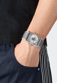 Riešas su ranka dryžuotų džinsų kišenėje, dėvintis stačiakampį sidabrinį laikrodį su pilku pintu dirželiu ir minimalistiniu apvaliu ciferblatu.