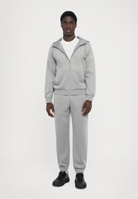 Grå træningssæt lavet af blødt stof, med en zip-op hoodie og elastiske manchetter. Båret med sorte snørebåndssko.