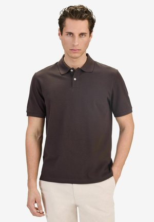 Man wearing a dark brown short-sleeve polo shirt and light beige pants, standing with a neutral expression and one hand in pocket.