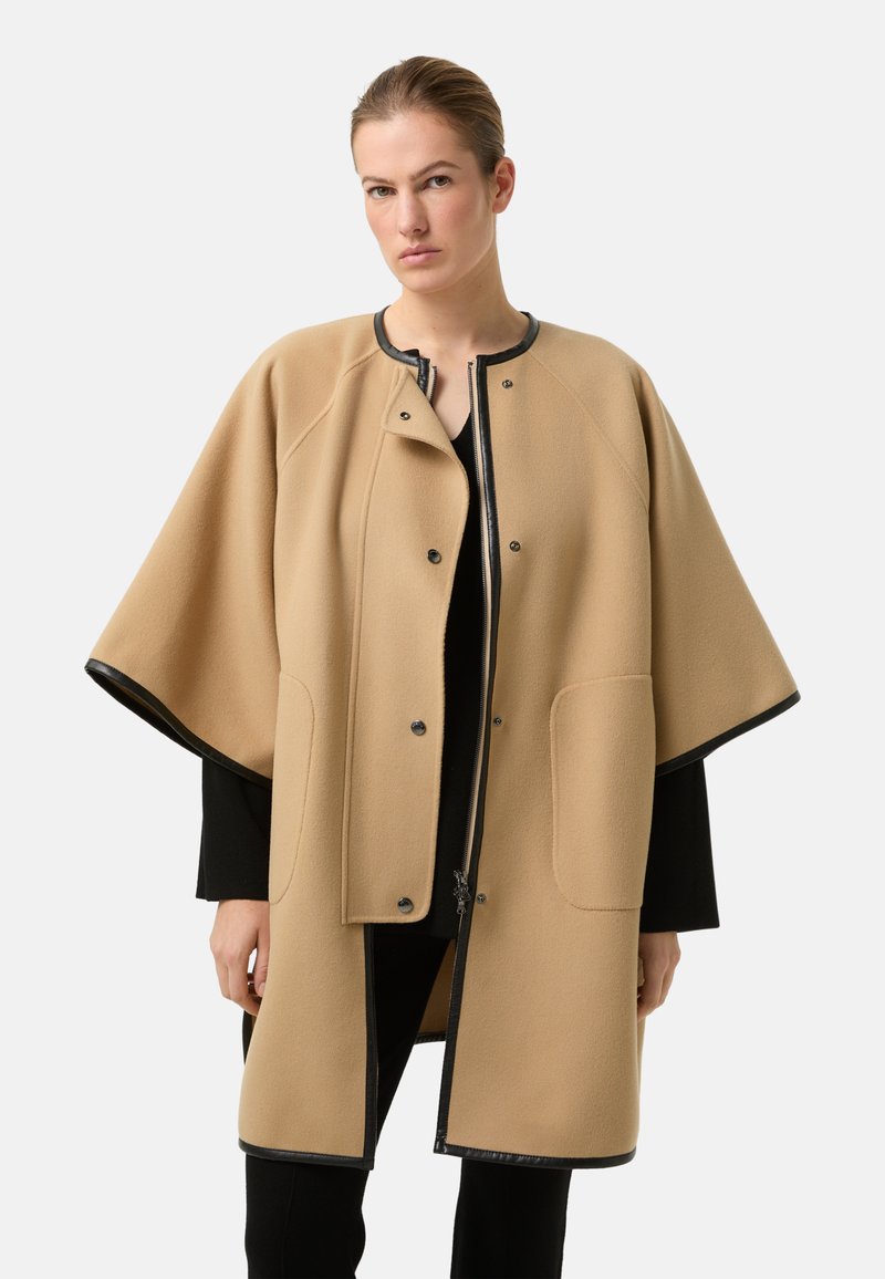 Beige uldblandet kappe med sort kant og trykknapper. Har en bred silhuet, to forlommer og en lynlåsåbning.