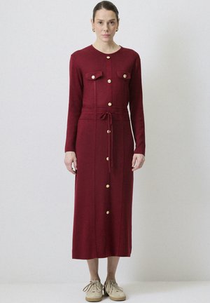 Robe longue bordeaux à manches longues avec un devant à boutons, deux poches poitrine, taille cintrée avec cordon de serrage et boutons dorés. Baskets portées.