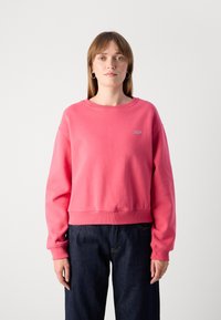 Pink cropped sweatshirt with long sleeves, a round neckline, and ribbed cuffs. Features a small logo on the chest, paired with dark jeans.