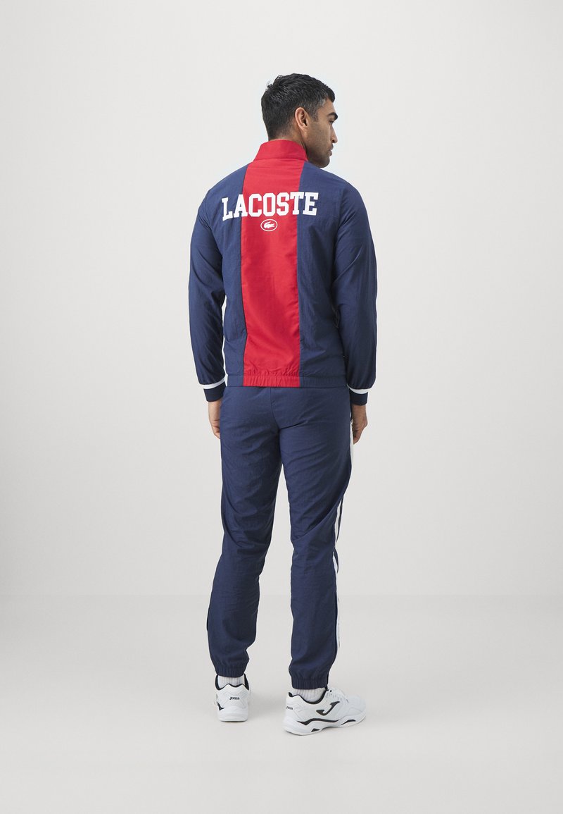 Ensemble de survêtement bleu marine et rouge. La veste présente le logo "LACOSTE" blanc au dos. Le pantalon a des accents rayés et des poignets élastiques. Le matériau semble léger.