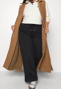 Lång kappa i camel med silverdetaljer, lagd över en vit fleecepullover och svarta joggers. Modellen bär silverfärgade sandaler.