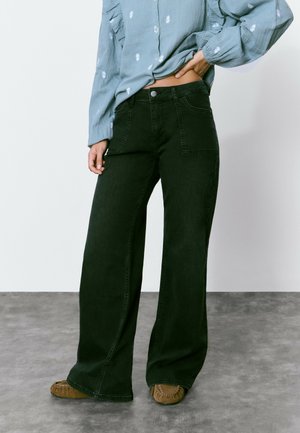 Jeans noirs à jambes larges avec une taille haute, comprenant deux poches avant et une texture subtile. Associés à une blouse bleu clair et des chaussures marron.