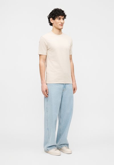 Νεαρός άνδρας στέκεται με το πρόσωπο πρ ος τα εμπρός, φοράει μπεζ κοντομάνικο πουκάμισο, ανοιχτό μπλε τζιν με φαρδιά πόδια και μπεζ αθλητικά παπούτσια, μπροστά σε απλό λευκό φόντο.