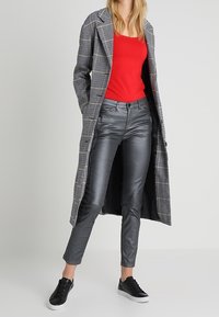 Cappotto lungo in motivo houndstooth, canotta rossa, pantaloni in pelle grigia lucida, sneakers nere. Il cappotto presenta un colletto a revers e una chiusura aperta.