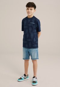 Tamsūs marškiniai su laiv�ų raštu šviesiai mėlyna spalva, derinami su šviesiai džinsiniais šortais. Avalynė - juodi ir turkio spalvos sportbačiai.