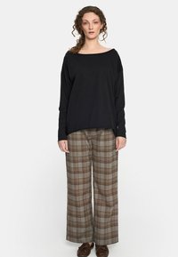 Kobieta z kręconymi włosami ma na sobie czarną bluzkę z długim rękawem, szerokie brązowe spodnie w kratę i brązowe loafersy, stojąc na tle jasnego, jednolitego tła.