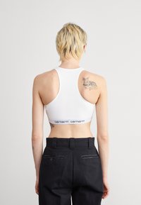 Crop top de sport blanc avec un dos nageur, présentant une bande logo noire. Associé à un pantalon noir taille haute.
