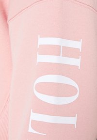 Roze sweatshirt gemaakt van zachte stof, met grote witte tekst "HOL" op de mouw. Het ontwerp is eenvoudig met een gladde textuur.