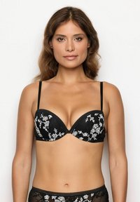 Donna che indossa un reggiseno nero di pizzo con ricami floreali bianchi, in piedi contro uno sfondo bianco semplice, rivolta in avanti.