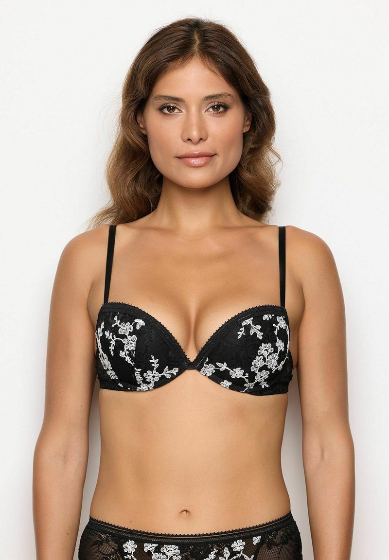 Donna che indossa un reggiseno nero di pizzo con ricami floreali bianchi, in piedi contro uno sfondo bianco semplice, rivolta in avanti.