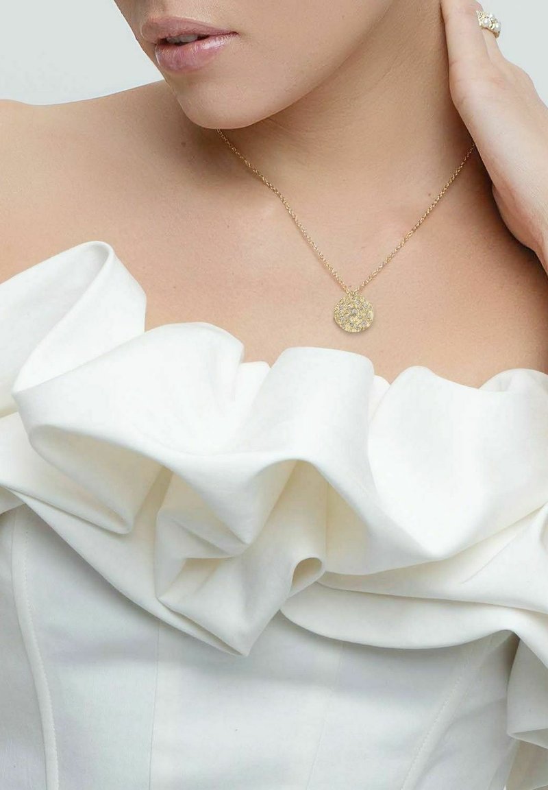 Guldig halsband med en texturerad rund pendant, bärs med en vit utan axelbands topp med framträdande volanger och slät tyg.