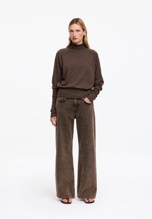 Femme portant un pull à col roulé marron et un jean marron à jambes larges, debout avec une main dans la poche sur fond blanc.