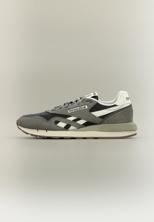 Šedá a čierna teniska značky Reebok s bielym pruhovaným dizajnom a logom, sivými šnúrkami a polstrovanou podrážkou na neutrálnej pozadí.