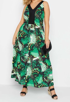Femme portant une robe maxi verte sans manches imprimée papillons avec un détail en V noir, tenant une pochette noire et portant des sandales compensées noires.