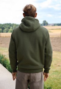 Felpa con cappuccio verde oliva realizzata in morbido tessuto, con una vestibilità rilassata, polsini e orlo a costine, indossata con pantaloni chiari.