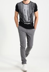 Černé tričko s grafikou městského panorama, spárované se světle šedými kalhotami. Bílé tenisky dotvářejí outfit, zdůrazňující ležérní styl.
