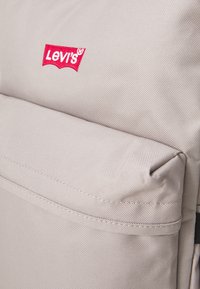 Šedý látkový ruksak s výrazným červeným logom Levi's. Materiál má textúrovaný povrch a obsahuje predné vrecko so švovými detailmi.