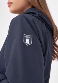 Marineblå jacka i glatt tyg, med huva och en texturerad lapp med ett slott design på vänster axel.