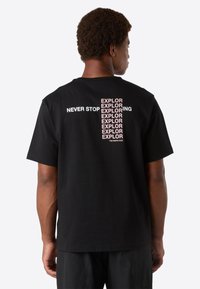 Man med svart T-shirt där texten "NEVER STOP EXPLORING" upprepas vertikalt i rosa och vitt på ryggen, står framför en enfärgad bakgrund.