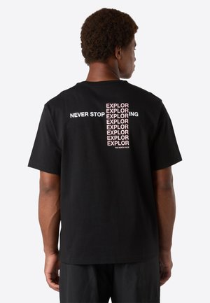 Un homme portant un T-shirt noir avec le texte "NE JAMAISE ARRETER D'EXPLORER" répété verticalement en rose et blanc dans le dos, se tenant devant un fond uni.