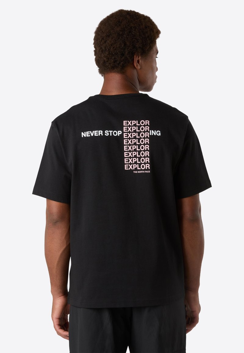 Man med svart T-shirt där texten "NEVER STOP EXPLORING" upprepas vertikalt i rosa och vitt på ryggen, står framför en enfärgad bakgrund.