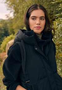Schwarze, gepolsterte Weste mit hohem Kragen, ausgestattet mit einem strukturierten schwarzen Fleece-Futter und Reißverschluss-Taschen, über einem dunklen Thermo-Oberteil getragen.