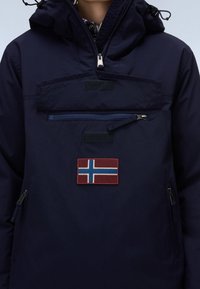 Person trägt eine dunkelblaue Kapuzenjacke mit einer norwegischen Flaggen-Aufnäher und einer Reißverschlusstasche vorne auf der Brust.