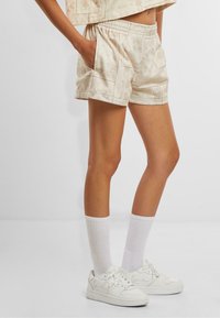 Beige gemusterte Shorts mit elastischem Bund, seitlichen Taschen und lockerer Passform, kombiniert mit weißen Kniestrümpfen und weißen Sneakern.